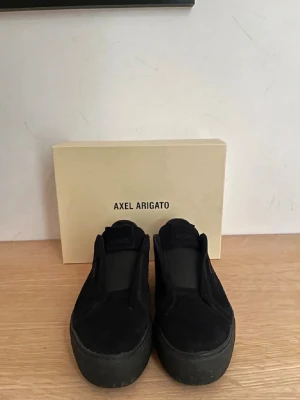 Svarta slip-on sneakers Axel Arigato - Stilrena svarta slip-on sneakers från Axel Arigato i mocka med diskret logga på sidan. Skorna har en chunky sula och minimalistisk design som passar till det mesta. Insidan är fodrad i läder för extra komfort.