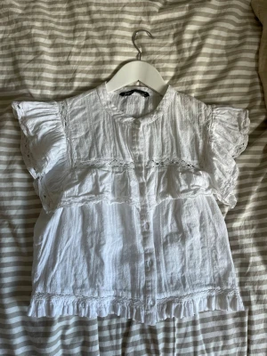 Vit topp Zara med volanger och spetsdetaljer - Söt vit blus med volanger på ärmar och nederkant samt fina spetsdetaljer. Blusen har knappar framtill och en rak passform. Tillverkad i ett lätt, luftigt tyg. Storlek L men sitter som en S