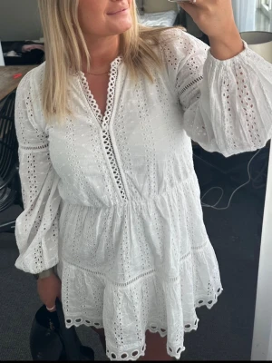 Vit broderad kortklänning med volang - Söt vit kortklänning med broderade detaljer och volangkant. Klänningen har v-ringning med spets, långa puffärmar och markerad midja. Perfekt för sommarens alla tillfällen och ger en romantisk känsla.
