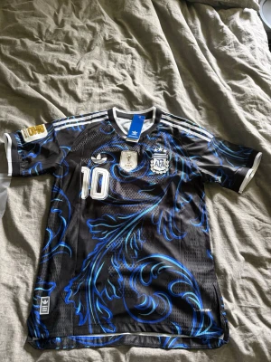 Argentina Messi tröja svart/blå Adidas - Säljer en svart Argentina fotbollströja med blått mönster, nummer 10 och Messi på ryggen. Tröjan har vita detaljer, officiella klubbmärken och är tillverkad i lätt, ventilerande material. Perfekt för dig som älskar fotboll och vill sticka ut på planen. Tröjan är aldrig använd därför 10/10 i skick. Vid frågor hör av er! Messi GOAT 