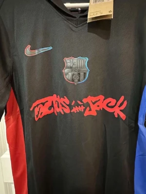 Svart FC Barcelona x Nike matchtröja - Snygg svart matchtröja från Nike i samarbete med FC Barcelona och Cactus Jack. Tröjan har röda och blå detaljer på sidorna, klubbmärke på bröstet och grafisk text framtill. Tillverkad i lätt och ventilerande material, perfekt för fotboll eller streetwear.