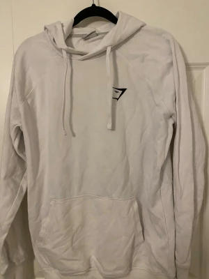 Vit hoodie från Gymshark med huva - Säljer en vit hoodie från Gymshark med klassisk huva och dragsnören. Hoodien har en känguruficka framtill och Gymshark-loggan broderad på bröstet. Tillverkad i mjukt material och har ribbade muddar vid ärmslut och nederkant. Perfekt för en avslappnad stil.