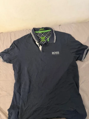 Hugo boss piké  - Snygg mörkblå pikétröja från BOSS med vit logga på bröstet och baksidan. Tröjan har klassisk krage, knappar framtill och vita ränder på krage och ärmslut. Perfekt för en stilren look.