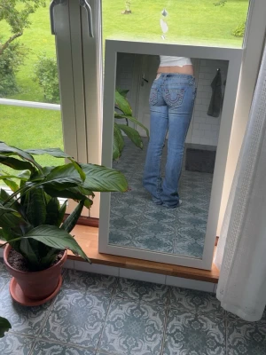 True religion jeans - Säljer ett par ljusblå bootcut jeans med snygga broderade detaljer på bakfickorna. Jeansen har låg midja och klassisk femficksdesign. Perfekta för dig som gillar en retro och avslappnad stil.