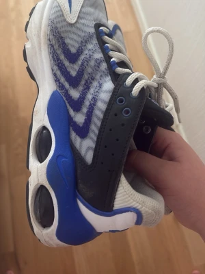 Nike Air Max Plus blå och vita sneakers - Nike Air Max Plus sneakers i vitt, blått och svart med tydliga vågmönster på ovandelen. Skorna har vita snören, svarta detaljer och blå accenter på sidorna samt synlig Air Max-dämpning i sulan. Ovandelen är i mesh och syntetmaterial för bra andningsförmåga.