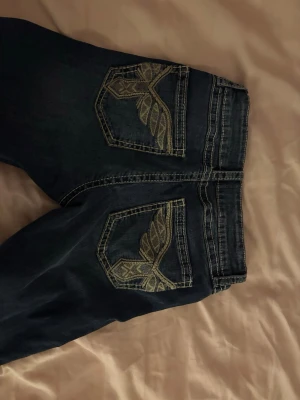 Blå bootcut jeans med broderade fickor - Säljer ett par blå bootcut jeans med snygga broderade detaljer i guld på bakfickorna. Jeansen har klassisk femficksmodell och kontrastsömmar. Perfekta för dig som vill ha en unik touch på din denimstil. Är storlek 170 men passar för dom som har S