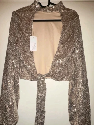 Glittrig silvrig knytblus från New Trend - Supersnygg glittrig silvrig blus från New Trend med långa ärmar och knyt framtill. Blusen har hög krage och är croppad, perfekt för att sticka ut. Materialet är glittrigt och skimrande, vilket ger en riktigt festlig vibe.
