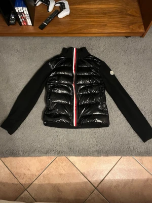 Moncler cardigan - Snygg svart dunjacka från Moncler med glansig front och stickade svarta ärmar. Jackan har två dragkedjeförsedda fickor och en röd-vit rand längs dragkedjan. Säljer då den ej passar mig, Scanningen på själva moncler märket funkar ej. Snabb affär