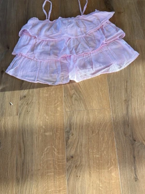 Ljusrosa volangig linnetopp - Supersöt ljusrosa linnetopp med tunna axelband och flera lager volanger. Den är luftig och perfekt för varma dagar, med en flowy och feminin look. Materialet känns lätt och mjukt mot huden.
