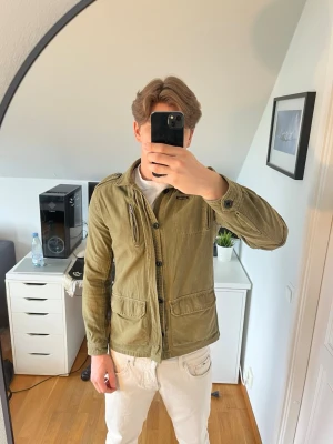Olivgrön utilityjacka med fickor - Snygg olivgrön utilityjacka med flera praktiska fickor framtill och dragkedja och knappar.  Hör gärna av dig vid frågor!
