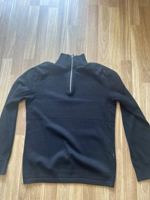 Svart stickad tröja Selected Homme - Svart stickad tröja från Selected Homme med halv dragkedja vid halsen och diskret strukturmönster över bröstet och ärmarna. Långärmad modell i mjukt material, perfekt för lager på lager och stilren look.