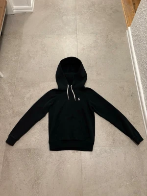 Polo Ralph Lauren hoodie  - Säljer en svart hoodie från Polo Ralph Lauren med  vita snören och en vit broderad logga vid bröstet och har ett mjukt material som känns riktigt skönt. Perfekt för dig som gillar stilrena och tidlösa plagg. Storleken är xxs i dam. Nypris 2195kr! Kan användas på både killar och tjejer. Även ett väldigt litet hål nere vid högra midjans baksida på tröjan, syns knappt med tanke på att tröjan är svart och hålet är några millimeter stort. Hör av er!
