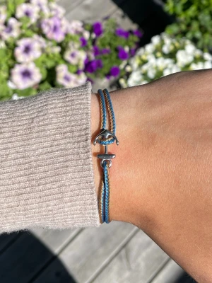 Blått och brunt armband med ankare - Snyggt armband med dubbla snören i blått och brunt. Armbandet har ett silverfärgat ankare som lås och är justerbart för att passa olika handleder. Perfekt accessoar för en avslappnad och trendig stil.