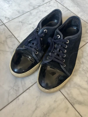 Mörkblå sneakers med lackad tå - Säljer mina Lanvin-skor i fint använt skick. Skorna är väl omhändertagna och fungerar precis som de ska. De har normala tecken på användning men är fortfarande väldigt snygga och passar till det mesta.  Det finns en mindre defekt på vänstra skon, vilket syns på sista bilden, så kolla gärna bilderna noggrant innan köp.  Ingen box eller kvitto finns kvar.  Priset är inte hugget i sten, så skicka gärna ett bud vid seriöst intresse. 