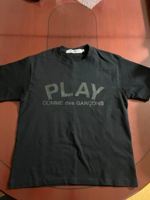 Svart PLAY t-shirt Comme des Garçons - Säljer en svart t-shirt från Comme des Garçons PLAY med stor logga tryckt på bröstet och ryggen. Klassisk rund hals och korta ärmar. Tillverkad i mjuk bomull för skön känsla och enkel stil.