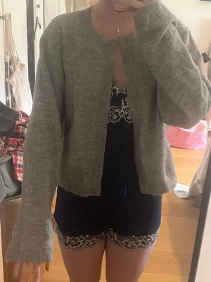 Grå kofta från H&M Divided - Mysig gråmelerad kofta från H&M Divided med rund halsringning och knappar framtill. Koftan har långa ärmar och ribbade muddar vid ärmslut och nederkant. Perfekt att slänga över en topp för en chill look.