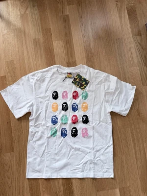 Vit BAPE t-shirt med färgglatt tryck - Säljer en vit t-shirt från BAPE med ikoniska ape-heads i flera färger på framsidan. T-shirten har rund hals, korta ärmar och en loose passform. Perfekt för dig som gillar streetwear och vill sticka ut med ett unikt tryck.
