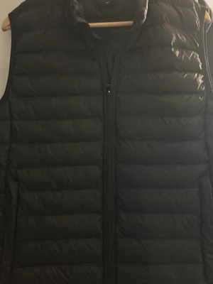 Svart dunväst med dragkedja - Stilren svart dunväst med horisontella sömmar och hög krage. Västen har en smidig dragkedja framtill och två sidofickor. Perfekt att slänga över en hoodie eller tröja för extra värme. Materialet är lätt och quiltat för en modern look.