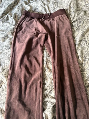 Bruna mjukisbyxor från Juicy Couture - Säljer ett par bruna mjukisbyxor från Juicy Couture i mjuk velour. Byxorna har bred resår i midjan med dragsko, två fickor fram och broderad JC-logga på ena benet. Perfekta för en chill och avslappnad stil.