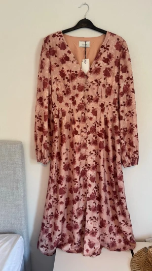 Blommig långklänning från Neo Noir - Säljer en otrolig snygg långklänning från Neo Noir i ljusrosa satin med vinröda blommor. Klänningen har v-ringning, långa ärmar med lätt puff och knappar framtill. Perfekt för sommarens alla tillfällen och ger en elegant look. Nypris 999kr