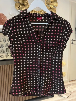 Brun blus med stjärnmönster  - Snygg brun blus från H&M med vita och rosa stjärnor över hela tyget i storlek S. Blusen har korta ärmar, krage och knäppning framtill samt en liten bröstficka. Materialet känns lätt och luftigt, perfekt för en trendig look. Axelbredd: 36 cm . Längd: 61 cm. Byst: 44 cm. 