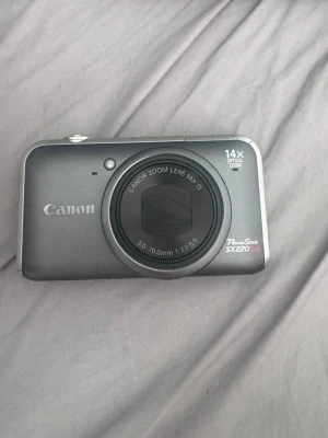 Canon PowerShot SX220 HS kompaktkamera - Snygg digitalkamera från Canon, modell PowerShot SX220 HS. Kameran är mörkgrå med silverdetaljer, har 14x optisk zoom och 12,1 megapixlar. Utrustad med bildstabilisering och ett smidigt kompakt format, perfekt för att ta med överallt.