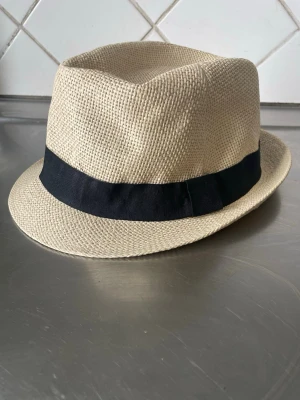 Beige fedora med svart band - Snygg beige fedora-hatt i flätad strådesign med ett brett svart band runt kullen. Klassisk form med något uppvikt brätte och markerad kulle. Perfekt accessoar för en trendig och avslappnad look.