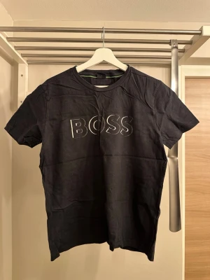 Svart BOSS t-shirt med tryck - Snygg svart t-shirt från BOSS med stort vitt logotryck på bröstet. Klassisk rund halsringning och korta ärmar. Tillverkad i mjukt bomullsmaterial som känns skönt mot huden. Perfekt för en clean och stilren look.