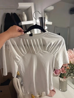 Vit ribbad t-shirt med rynkade detaljer - Snygg vit t-shirt med korta ärmar och unika rynkade detaljer över bröstet. Tröjan har en ribbad sektion framtill och en klassisk rund halsringning. Perfekt för dig som gillar stilrena men ändå speciella plagg.