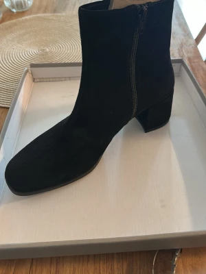 Svarta stövletter från Vagabond  - Snygga svarta stövletter med mellanhög blockklack och dragkedja på sidan. Skorna har en stilren design med rundad tå och är tillverkade i mockaliknande material. Perfekta för att lyfta din outfit med en trendig touch.
