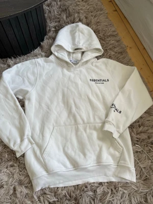 Vit Essentials hoodie  - Säljer en clean vit hoodie från Essentials i storlek XXL. Hoodien har huva sitter som L, känguruficka och tryck med 'ESSENTIALS Fear of God' på bröstet och ärmen. Mjuk insida och ribbade muddar. Perfekt för dig som gillar oversized streetwear vibes.