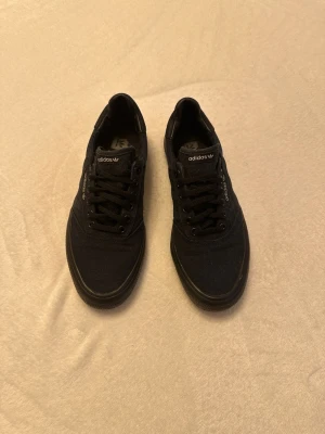 Svarta Adidas sneakers i canvas - Snygga svarta sneakers från Adidas med klassisk låg profil och svart sula. Skorna har ovandel i canvas, svarta skosnören och diskret Adidas-logga på plösen och sidan. Perfekta för en clean och stilren look.
