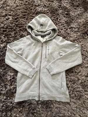Ljusgrå zip-hoodie från Sail Racing - Snygg ljusgrå zip-hoodie från Sail Racing. Tröjan har en bröstficka med zip. Klassisk huva med broderad logga och mjukt material. Perfekt för en avslappnad och sportig stil. Tröjan är i barnstorlek 170 men jag skulle rekommendera den om du är lite kortare.
