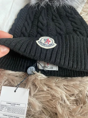 Svart mössa med äkta  pälsboll - Snygg svart stickad Moncler mössa med bred uppvikt kant och klassisk Moncler-logga framtill. Mössan har en stor, fluffig pälsboll i grått och svart på toppen. Kabelstickat mönster ger extra stilpoäng. Perfekt accessoar för kalla dagar.