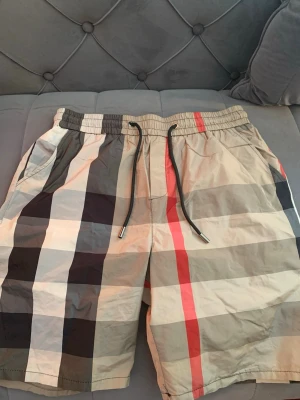 Rutig shorts med snörning - Snygga rutiga shorts med breda beige, svarta, vita och röda ränder. Shortsen har elastisk midja med svart snörning och sidofickor samt en bakficka. Perfekta för sommaren och har en avslappnad passform. Shortsen är helt nya, man kan både basa och hänga runt med de. De är L men passar oså S och M