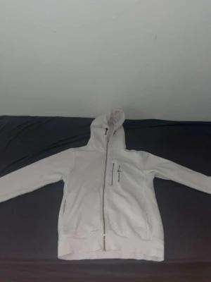 Vit hoodie från Sail Racing - Vit hoodie med dragkedja från Sail Racing. Tryck med logga på bröstet och stor text 'SAIL RACING EST. 1977' på ryggen. Utrustad med huva och två fickor framtill. Tillverkad i mjukt material, perfekt för en avslappnad stil.