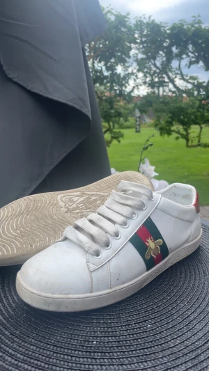 Vita Gucci sneakers med bi-detalj - Säljer ett par vita sneakers från Gucci med klassiska gröna och röda ränder på sidan och en guldfärgad bi-detalj. Skorna har vita snören och låg profil. Tillverkade i läder med gummisula och snygga detaljer som sticker ut. Priset är ej hugget i en sten