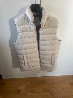 Beige dunväst från Reserved - Stilren beige dunväst från Reserved med hög krage och dragkedja framtill. Västen har två fickor på sidorna och quiltad design som ger en puffig look. Perfekt för lager-på-lager och enkel att matcha med olika outfits.