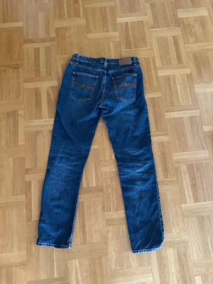 Blå jeans från Nudie Jeans - Säljer ett par klassiska blå jeans från Nudie Jeans med raka ben och snygga slitningar. Jeansen har två bakfickor med Nudie Jeans karakteristiska sömmar och är gjorda i mjuk denim. Perfekta för dig som gillar en tidlös och avslappnad stil.