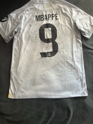 Real Madrid Mbappé matchtröja - Säljer en vit Real Madrid matchtröja med Mbappe #9 på ryggen. Tröjan har svarta och gula detaljer, klubbmärke på bröstet och sponsortryck 'Emirates Fly Better' framtill. Tillverkad i lätt och ventilerande material, perfekt för fotbollsträning eller match. Notera att numret är fel för året.