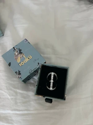 Silverfärgad statementring Ioaku - Säljer en unik justerbar silverfärgad statementring från Ioaku som jag aldrig har använt. Ringen har en öppen design med gnistrande stenar längs hela framsidan som ger en lyxig känsla. 