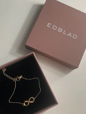 Guldarmband med infinitysymbol - Guldarmband från EDBLAD med en stilren infinitysymbol dekorerad med små gnistrande stenar. Tyvärr har några av stenarna lossnat därav varför jag säljer det. Armbandet är i rostfritt stål.