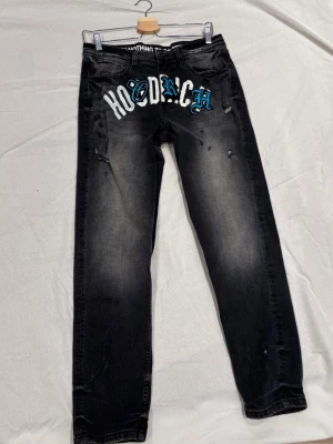 Svarta jeans från Hoodrich med tryck - Säljer ett par svarta jeans från Hoodrich med stort vitt och blått tryck över framsidan. Jeansen har en tvättad look, raka ben och slitna detaljer för en streetwear-känsla. Perfekta för dig som gillar en edgy stil.