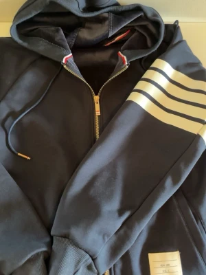 Svart hoodie med vita ränder Thom Brown  - Snygg svart hoodie från New Yoak med dragkedja och huva. Ärmen har fyra vita ränder som ger en sportig vibe. Dragsko i huvan med metalländar och ribbade muddar. Märkeslapp på magen. Perfekt för dig som gillar streetwear och enkel stil. Thom Brown 