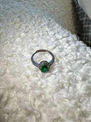Silverring med grön sten - Snygg silverfärgad ring med en stor rund grön sten i mitten och små klara stenar runt om. Ringen har en klassisk design och gnistrar fint i ljuset. Perfekt accessoar för att lyfta din stil.
