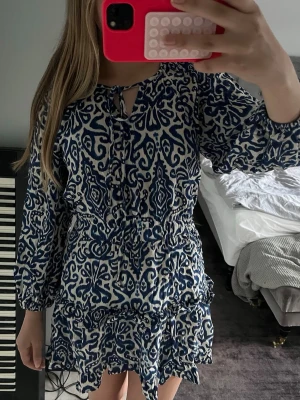 Blåvit mönstrad kortklänning Zara - Kortklänning från Zara i blått och vitt med unikt grafiskt mönster. Klänningen har långa ärmar med resår vid handlederna, volangdetaljer vid kjolen och knytning vid halsen. Perfekt för dig som gillar trendiga och bekväma plagg med snygg passform.