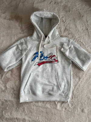 Grå hoodie Polo Ralph Lauren - Snygg grå hoodie från Polo Ralph Lauren med korta ärmar och stor huva. Framtill finns en stor logga i blått, vitt och rött med stjärnor och texten 'Polo Ralph Lauren'. Hoodien har känguruficka och vita dragsnören. Perfekt för en avslappnad streetwear-look. Strl:M men passar S. 100%äkta