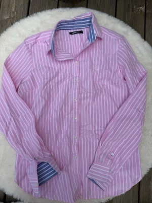 Rosa och vit randig skjorta Gina Tricot (S) - Säljer en rosa och vit randig skjorta från Gina Tricot i storlek 36. Skjortan har blå- och vitrandiga detaljer på insidan av kragen och manschetterna, klassisk krage, långa ärmar och knäppning framtill. Perfekt för en trendig och avslappnad look.