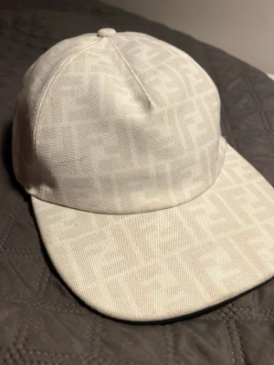 Vit keps från Fendi med logomönster - Säljer en vit keps från Fendi med det klassiska FF-logomönstret ton-i-ton över hela kepsen. Kepsen har böjd skärm och är tillverkad i ett slitstarkt tygmaterial. Perfekt för dig som vill ha en stilren accessoar med exklusiv känsla.