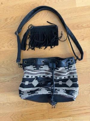 Svart och vit axelremsväska med fransar - Säljer en svart och vit axelremsväska med etniskt mönster och detaljer i konstläder. Väskan har dragskostängning med fransar och matchande liten clutch med fransar. Perfekt för dig som gillar boho-stil och vill ha något unikt till din outfit.
